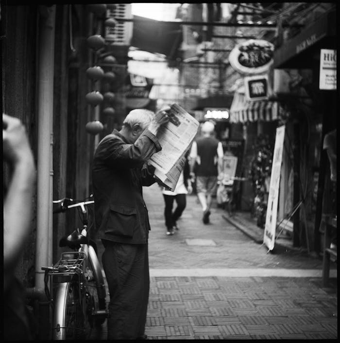 Elderly man reading a newspaper in a narrow city alleyway, a candid black and white street scene.